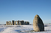 Stonehenge defined by early morning snow with a blue sky and the Heel Stone off to the right of the stone circle; Wiltshire, England, United Kingdom