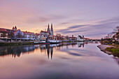 Outlook over the Danube River with the old, 12th Century Stone Bridge in the distance and the Gothic St Peter's Cathedral from the Marc?-Aurel-shore in the Old Town of Regensburg at sunset with a purple hue; Regensburg, Bavaia, Germany