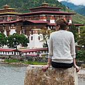 Eine Frau sitzt auf einem Steinvorsprung mit Blick auf den Punakha Dzong; Distrikt Punakha Bhutan
