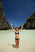 Eine Touristin mit Sonnenhut und Bikini steht mit erhobenen Armen im klaren Wasser der Insel Matinloc bei El Nido und Corong Corong; Bacuit Archipel; Palawan Philippinen.