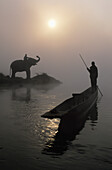 Ein Elefant mit seinem Mahout steht am Rande des Rapti-Flusses in der Nähe des Sauraha- und Chitwan-Nationalparks, während ein Mann sein Einbaum-Kanu den Fluss entlang schiebt; Nepal.