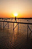 A Man Walks On A Pier With Boats Moored In The Water At Sunset Near Faro; Algarve Portugal