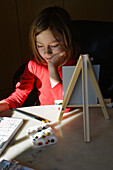 Young Boy with Shoulder-Length Hair sitting at Table with Art Supplies and Small Easel