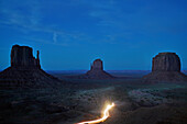 Monument Valley, Utah, Usa Autoscheinwerfer leuchten am Fuße der linken und rechten Mitten und Merrick Butte im Monument Valley an der Grenze zwischen den Bundesstaaten Utah und Arizona.