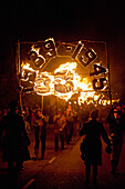 Menschen tragen ein großes Banner vor 31 brennenden Mohnblumen in der Nacht des Freudenfeuers zum Gedenken an die 31 in den Weltkriegen gefallenen Dorfbewohner, East Hoathly, East Sussex, Großbritannien