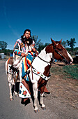 Ein Crow-Indianer reitet auf einem Pferd und ist während der Parade auf der jährlichen Crow-Messe (August), dem größten indianischen Fest in den USA, in traditionelle Insignien gekleidet. Die Crow (Apsaaloke, Apsaroke oder Absaroke) leben im Crow-Reservat (Montana, USA).