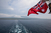 Red Australian Ensign flag flying from the stern of an expedition boat sailing around Karkar Island in the Bismarck Sea off the north coast of Papua New Guinea; Karkar Island, Madang, Papua New Guinea