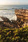 Eine Person steht auf einem zerklüfteten Bergrücken und blickt auf das Meer und die an die Küste rollenden Wellen; Praia do Guincho, Cascais, Portugal