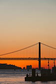Silhouette of the 25 de Abril Bridge crossing the Tagus River, connecting Lisbon and Almada at sunset; Lisbon, Estremadura, Portugal