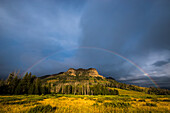 Regenbogen über dem Trident Ridge unter grau-blauem Himmel mit dem sonnenbeschienenen goldenen Gras der Ebenen in The Thorofare im Upper Yellowstone River Valley, Yellowstone National Park; Wyoming, Vereinigte Staaten von Amerika