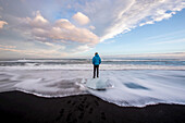 A man stands on washed up glacier ice on a black sand beach.