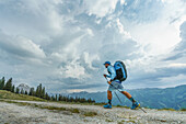 An athlete in the Redbull X Alps, a paragliding race, hikes with determination along the mountain trail to a summit to launch his paraglider under a stormy sky; Wagrain, Salzburgerland, Austria