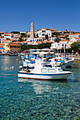 Nahaufnahme von Fischerbooten am Hafen von Emborio mit traditionellen Gebäuden und dem Uhrenturm im Stadtzentrum auf der Insel Chalki (Halki); Dodekanes-Inselgruppe, Griechenland