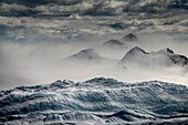 Der Eisberg im Vordergrund wirkt wie eine Bühne für den windgepeitschten Schnee, der an den Bergen der Iceberg Alley auf dem Weg zur westlichen Antarktischen Halbinsel vorbeifliegt; Antarktis