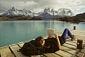 A woman relaxes on a dock while reading a book.; Torres del Paine National Park, Patagonia Chile.