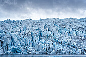 Close-up views taken from of the the Fjallsarlon Glacier Lagoon of the jagged blue ice shapes of the Fjallsjokull Glacier terminus which stands out against the grey, misty clouds, located at the south end of the famous Icelandic glacier Vatnajökull in Southern Iceland; South Iceland, Iceland