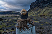 Nahaufnahme einer Frau mit blondem Haar, von hinten aufgenommen, die eine Wollmütze und einen isländischen Pullover trägt, während sie draußen in der Landschaft steht und die natürliche Schönheit der Berge und des stürmischen Himmels bewundert; Südisland, Island