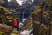 Frau steht und überblickt die Mulagljufur-Schlucht, ein Wanderparadies, und genießt die fantastische Aussicht auf einen Wasserfall und die moosbewachsenen Klippen; Vik, Südisland, Island
