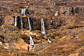Scenic view of a woman standing in front of a series of waterfalls flowing from the craggy cliffs of the East Fjords making her appear small against the vast landscape in front of her; East Iceland, Iceland