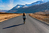 Woman walking down an open road towards Seyðisfjörður with a view of the snow-capped mountains and stark beauty of the East Fjords under a blue sky; East Iceland, Iceland