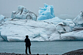 Silhouette of woman in a snowsuit standing on the shore, looking at the beautiful icebergs and amazing, blue ice-formations and shapes of the Jökulsárlón Glacier Lagoon located at the south end of the famous Icelandic glacier Vatnajökull in Southern Iceland; South Iceland, Iceland