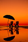 Dramatische Sonnenuntergangsszene mit zwei Stühlen und einem Sonnenschirm am Strand; Seaside, Florida, Vereinigte Staaten von Amerika