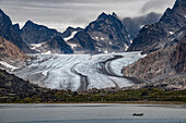 Blick auf einen Gletscher, der durch die Berggipfel an der Südspitze Grönlands fließt, mit der Silhouette eines vorbeifahrenden Bootes im grauen Wasser des Prins Christian Sund; Südgrönland, Grönland