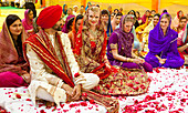 Braut und Bräutigam sitzen inmitten von Konfetti zwischen Anwesenden und Gästen; Ludhiana, Punjab, Indien