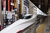 Japanischer Hochgeschwindigkeitszug wartet am Bahnhof mit Gebäuden dicht dahinter; Tokio, Japan