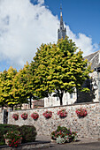Eine mit hängenden Blumentöpfen geschmückte Steinmauer und die Spitze einer Kirche im Hintergrund; Hargnies Ardennen Frankreich