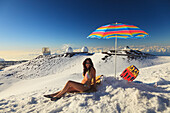 A Woman In Bikini Sitting In The Snow With Beach Umbrella With The View From Mauna Kea Observatories; Mauna Kea Hawaii United States Of America