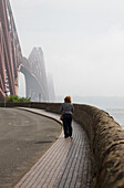 A Woman Walking On A Pathway On The Water's Edge Towards Forth Bridge; North Queensferry Scotland