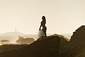 Silhouette of a female surfer holding her surfboard while standing on a rock at the coast; andalusia spain