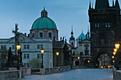 Tschechische Republik, Kreuz und Statue, Denkmal mit Laternenpfählen entlang des Weges, beleuchtet in der Dämmerung; Prag
