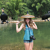 Ein Mädchen posiert mit einem einzigartigen Hut und einem Stock, der zwei Vögel entlang eines Flusses hält; Guilin, Guangxi, China