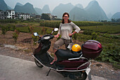 Eine Frau posiert mit ihrem Motorroller mit den spitzen Bergen im Hintergrund; Yangshuo, China