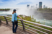 Eine Frau steht an einem Geländer mit Blick auf die Horseshoe Falls im Niagara Falls State Park im Bundesstaat New York, mit der Stadt Niagara Falls, Ontario im Hintergrund; Niagara Falls, New York, Vereinigte Staaten von Amerika