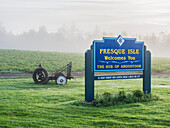 Welcome Sign To Presque Isle, Maine On A Foggy Summer Morning With Farm Equipment, A Potato Field And Dewy Grass Surrounding It; Presque Isle, Maine, United States Of America