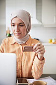 Lächelnde Frau mit Hidschab, die beim Online-Shopping eine Kreditkarte in der Hand hält