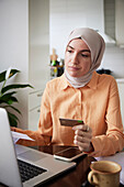 Lächelnde Frau mit Hidschab, die beim Online-Shopping eine Kreditkarte hält