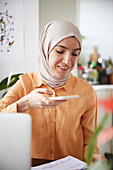 Smiling woman with hijab using cell phone and scanning bills