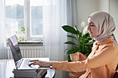Lächelnde Frau mit Hijab bei der Arbeit von zu Hause mit Laptop