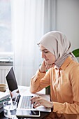 Lächelnde Frau mit Hijab, die von zu Hause aus arbeitet und einen Laptop benutzt