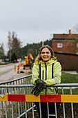 Lächelnde Straßenbauarbeiterin lehnt an einer Verkehrsschranke