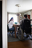 Mother and disabled teenage daughter in wheelchair sitting at kitchen table