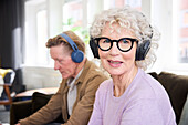 Lächelnde ältere Frau schaut in die Kamera, während sie Kopfhörer trägt, älterer Mann im Hintergrund