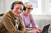 Lächelnder älterer Mann schaut in die Kamera, während er Kopfhörer trägt, ältere Frau im Hintergrund