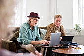Ältere Männer reden miteinander, während sie im Wohnzimmer sitzen und ein digitales Tablet und einen Laptop benutzen, um einen Podcast zu bearbeiten
