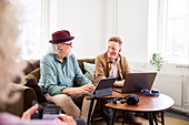 Ältere Männer sprechen miteinander, während sie im Wohnzimmer sitzen und ein digitales Tablet und einen Laptop benutzen, um einen Podcast zu bearbeiten
