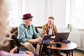 Senior men talking to each other while sitting in living room and using digital tablet and laptop to edit podcast